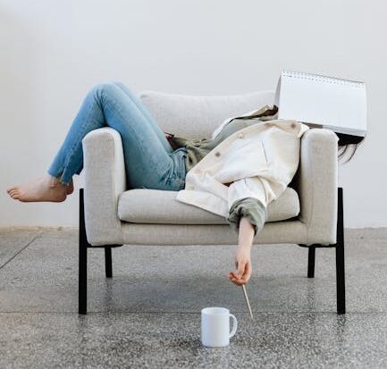 Tired woman in casual attire falls asleep on sofa with a notebook on face and pen in hand.